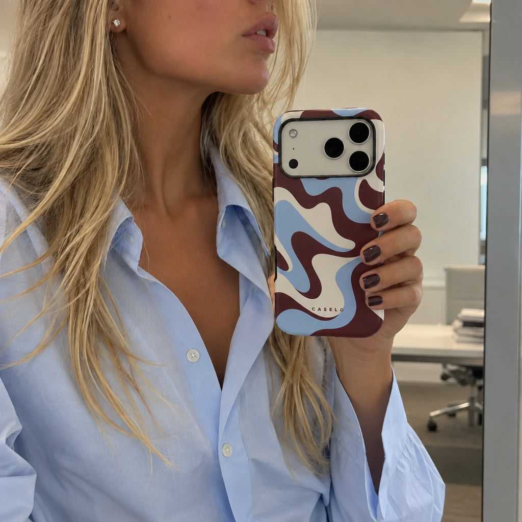 A woman in a light blue button-down shirt holds up a Samsung smartphone showing off a wavy abstract pattern phone case. The case features a maroon, light blue, and white design with the brand name 'SILKWORKS' printed in small white letters near the bottom.