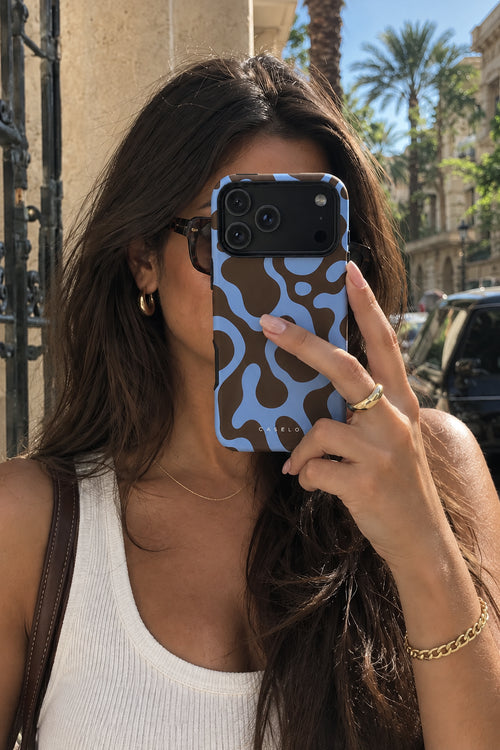 A woman taking a mirror selfie outdoors, holding a light blue and dark brown abstract patterned phone case in front of her face. The background shows a sunny street with palm trees, buildings, and parked cars.