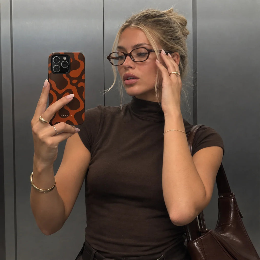 A woman takes a mirror selfie wearing a brown mock neck t-shirt and glasses, holding a phone with an orange and black patterned case. She is standing in front of a metallic gray wall.