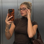 A woman takes a mirror selfie wearing a brown mock neck t-shirt and glasses, holding a phone with an orange and black patterned case. She is standing in front of a metallic gray wall.