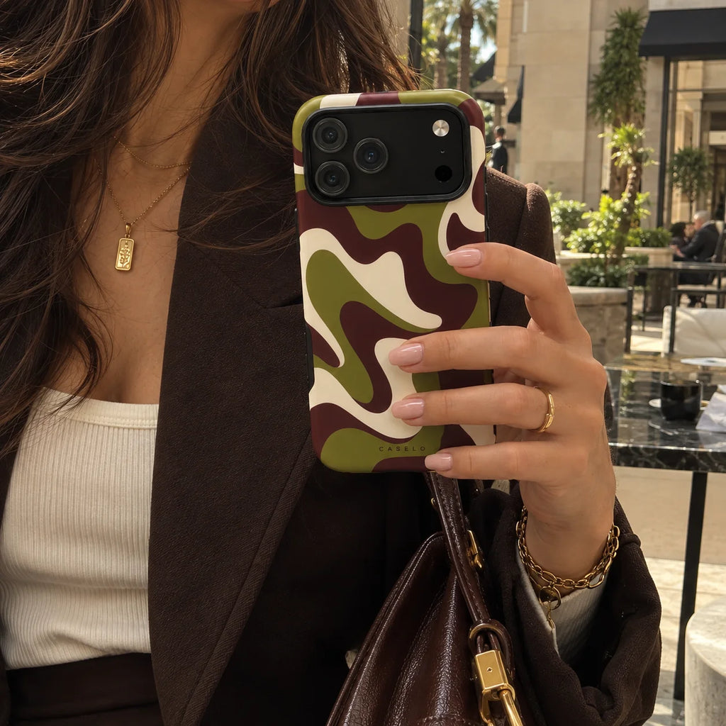 A close-up lifestyle shot of a person holding a smartphone with a green, brown, and beige wavy camouflage pattern phone case. The person is wearing a brown blazer, a white ribbed top, and a brown leather shoulder bag, with a cafe setting visible in the background.