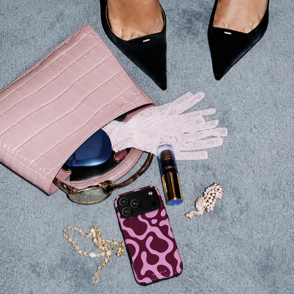 A styled flat-lay of a pink crocodile-embossed handbag on a textured gray surface, with a white lace glove, black pointed flats, a purple phone case, sunglasses, a tube of lipstick, a pearl bracelet, and a gold chain arranged around it.
