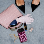 A styled flat-lay of a pink crocodile-embossed handbag on a textured gray surface, with a white lace glove, black pointed flats, a purple phone case, sunglasses, a tube of lipstick, a pearl bracelet, and a gold chain arranged around it.