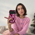 A woman in a pink knit cardigan holding a pink phone case with a wavy abstract pattern, taking a mirror selfie in a cozy indoor setting.