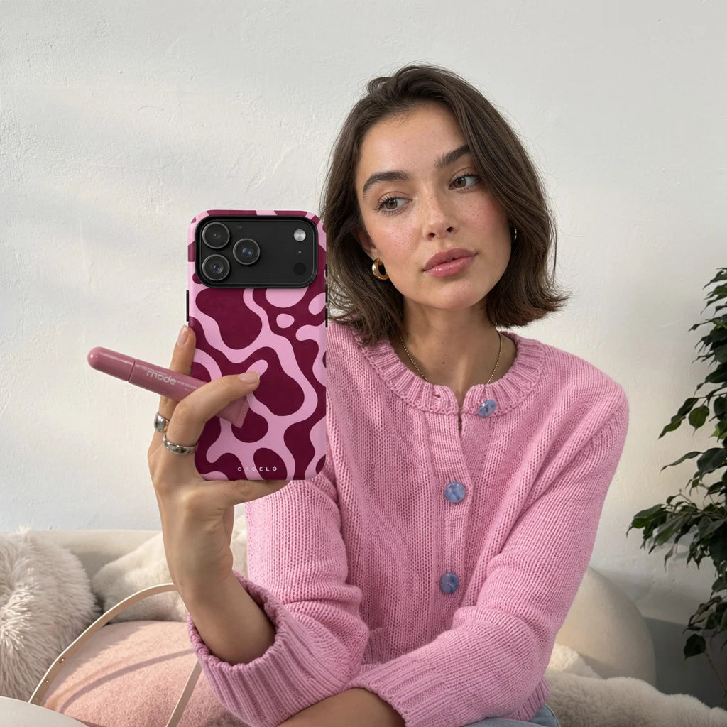 A woman in a pink knit cardigan holding a pink phone case with a wavy abstract pattern, taking a mirror selfie in a cozy indoor setting.